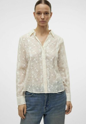 Blouse blanche à motifs floraux transparent, avec un col, une fermeture à boutons sur le devant et des manches longues, agrémentée d'une taille légèrement raccourcie.