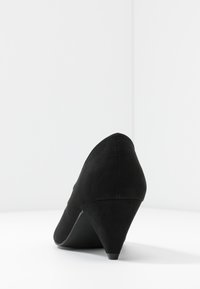 Chaussure à talon haut en daim noir avec un bout pointu et un talon modéré en forme de cône. Texture lisse, design simple, sans ornements visibles.