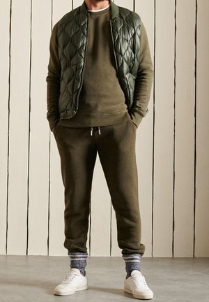 Veste matelassée vert olive portée sur un pull assorti, accompagnée d'un pantalon de jogging vert olive. Baskets blanches et chaussettes grises à motifs. Matériaux texturés.