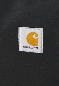 Étiquette en tissu noir avec une bordure blanche et un logo Carhartt jaune présentant un design en forme de C, affichant du texte rouge pour le nom de la marque.
