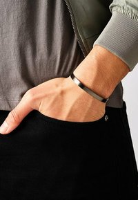 Bracelet en métal avec finition noire mate, présentant un design épuré et minimal. Porté sur un poignet, associé à une chemise grise et un pantalon noir.