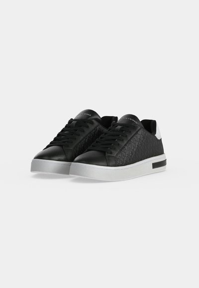 Une paire de baskets noires à lacets, avec des empiècements latéraux texturés, des semelles blanches et de petits détails blancs au talon.
