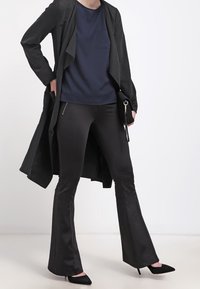 Svart trenchcoat över en marinblå topp, kombinerad med svarta utsvängda byxor och spetsiga klackar. Har en framficka och dragkedjeaccent.