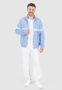 Giacca zip blu chiaro con strisce bianche, realizzata in tessuto traspirante. Abbinata a pantaloni bianchi e sneakers bianche, per un look sportivo.