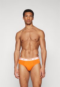 Oranžne Nike spodnjice z belim pasom, na katerem je logotip Nike. Tkanina je gladka in oprijeta, zasnovana za udobje in oporo.