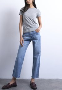 Γυναίκα που φοράει ανοιχτό γκρι κοντομάνικο πουκάμισο, τζιν παντελόνι υψηλής μέσης σε ανοιχτό μπλε χρώμα με ίσια γραμμή και σκουροκόκκινα μοκασίνια, στέκεται με το ένα χέ ρι στην τσέπη.