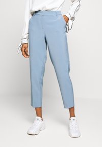 Person som bär ljusblå kapade byxor, vita sneakers och en vit blus med svarta detaljer knutna vid manschetterna, poserande med ena handen i fickan.