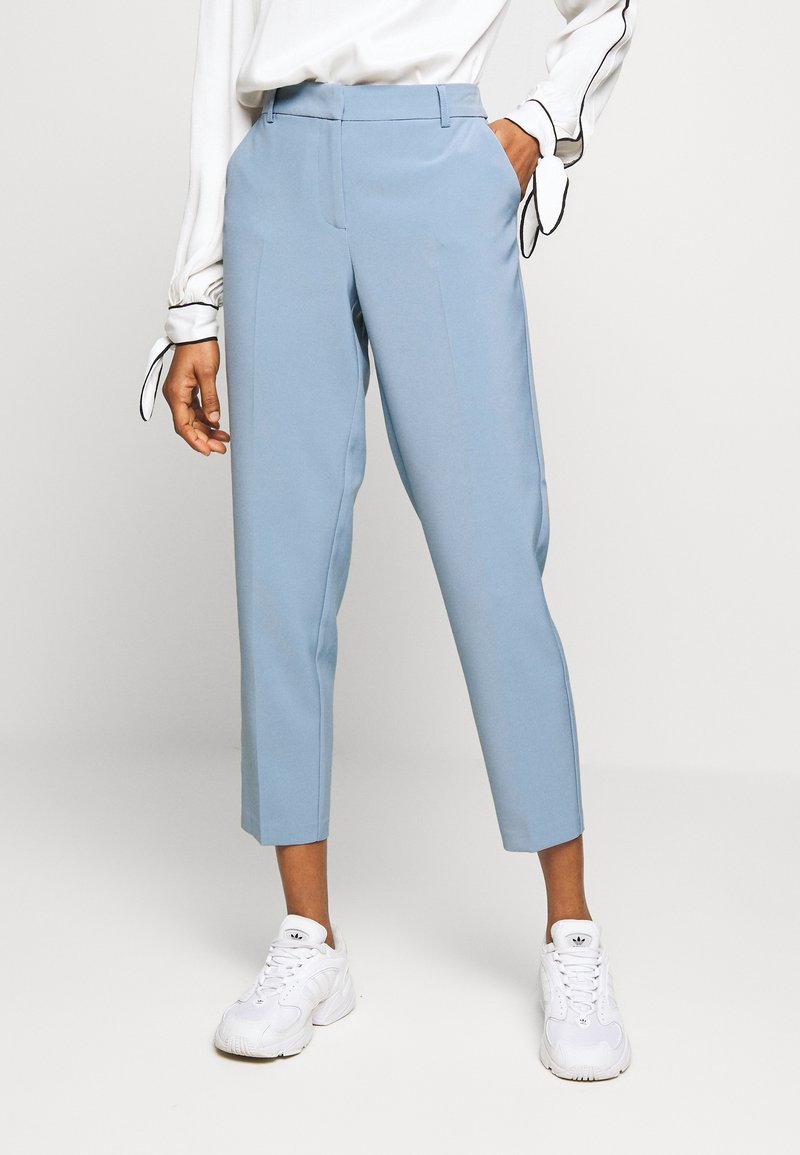 Person som bär ljusblå kapade byxor, vita sneakers och en vit blus med svarta detaljer knutna vid manschetterna, poserande med ena handen i fickan.