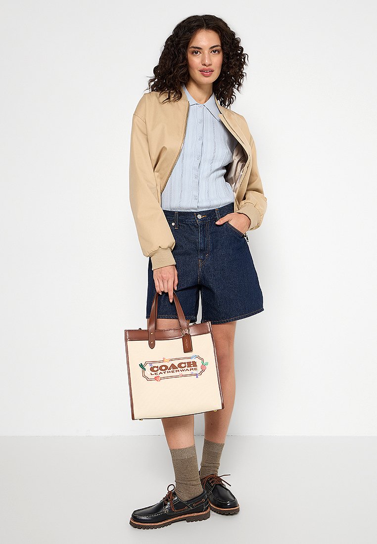 Chaqueta bomber beige, camisa de punto de cable azul clara, pantalones cortos de mezclilla oscuros, bolso de piel marrón con acentos florales, zapatos de cordones marrones y calcetines tobilleros beige.