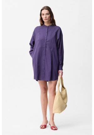 Donna che indossa un vestito viola largo con bottoni, tiene una borsa tote beige intrecciata e sandali rossi aperti sulle dita, in piedi su sfondo bianco.