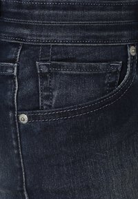 Mörkblå denimjeans har en bakficka, silverfärgade knappar och kontrasterande sömmar längs midjebandet och fickkanterna.
