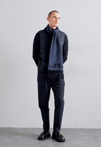 Un gilet matelassé noir porté sur une chemise noire à manches longues, associé à un pantalon sombre et une écharpe bleue à motif géométrique avec des franges.