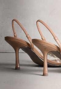 Paire de chaussures à talons hauts couleur camel avec fines brides tissées et talons pointus sur une surface grise, sur un fond neutre.