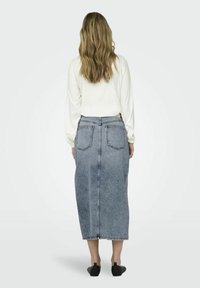 Falda de mezclilla en azul claro con dos bolsillos traseros, de longitud media hasta la pantorrilla y corte recto. Combinada con una blusa blanca de manga larga y zapatos planos negros.