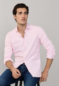 Jonge man zittend op een kruk, draagt een lichtroze overhemd met opgerolde mouwen en blauwe jeans, kijkt bedachtzaam weg.