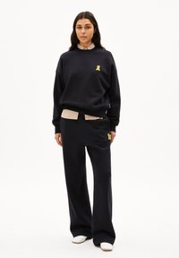 Marineblauer Sweatshirt mit einem Logo in Form eines gelben Ribbons, kombiniert mit marineblauen Weitbeinhosen, die einen gestreiften Bund und zwei Seitentaschen haben.