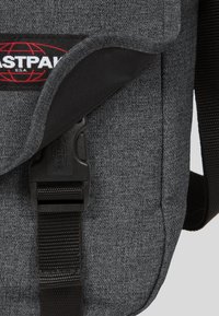 Borsa a tracolla in tessuto grigio con una tracolla nera, caratterizzata da un pattino frontale e una fibbia in plastica. Include un'etichetta con logo rosso e nero.