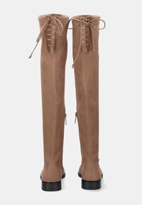 Botas de ante color beige hasta la rodilla con detalles de cordones en la parte trasera, textura suave y cierre de cremallera a los lados. Suelas planas negras.