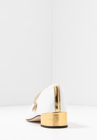 Scarpa bianca con punta tondeggiante e tacco basso in metallo dorato, caratterizzata da un design elegante e un dettaglio con bottone laterale. Superficie liscia.