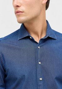 Denim shirt in donkerblauw met een puntkraag, knopenlijst aan de voorkant en witte knopen. De stof lijkt zacht met een licht gestructureerde afwerking.