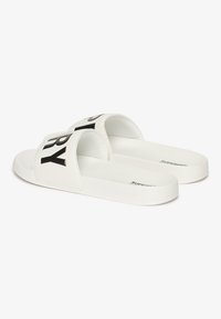 Hvide skydesandaler med en tekstureret rem, der har sort "SUPERDRY" skrift. Glat overflade, flad sål og minimalistisk design.