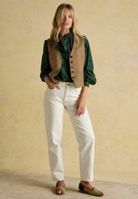 Braun karierter Weste über einer grünen Rüschbluse, kombiniert mit crèmefarbenen, weiten Hosen und mehrfarbigen, gemusterten Schuhen. Strukturierten Hintergrund.