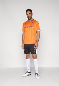 Tricou de fotbal portocaliu din plasă cu accente și logo mov. Pantaloni scurți negri cu detalii albe. Șosete albe și ghete negre de fotbal completează complet ținuta.