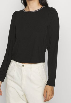 Persoon draagt een zwarte top met lange mouwen met een kralenversiering op de halslijn en een witte broek met hoge taille, staand tegen een effen achtergrond.