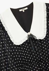Sort prikket kjole med en hvid flæsekrave, der har en perle-accent og sort bånd detalje ved halsudskæringen. Letvægtsstof.