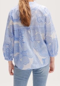 Blouse à fleurs bleu clair avec une coupe lâche, de longues manches bouffantes et un dos froncé. Le tissu a une texture semi-transparente et un imprimé complexe.