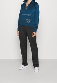 Blå zip-up sweatshirt med en metallisk logotyp, tillsammans med svarta byxor och vita sneakers. Tyget har en slät yta.