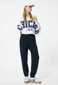 Vit kortärmad sweatshirt med "CHICAGO U.S." i marinblått, kombinerad med marinblå joggers. Modellen bär en vit keps och vita sneakers med marinblå detaljer.