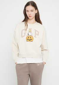 Εκρού φόρμα με στάμπα Snoopy και κολοκύθα, έντονα γράμματα "GAP" και ριμπ μανσέτες. Φορεμένη πάνω από λευκό πολυστρωματικό πουκάμισο.