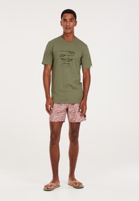 Camiseta verde oliva con estampado gráfico negro; bañador corto rosa con patrones geométricos; sandalias planas. Estilo simple y deportivo.