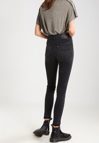 Žena oblečená v tmavých skinny džínech, olivově zeleném tričku a černých kotníkových botách, stojící zády k fotoaparátu na jednolitém pozadí.