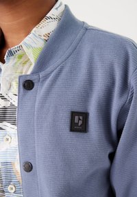 Cardigan azzurro chiaro con una texture a coste, caratterizzato da una toppa nera con logo, chiusura a bottoni e sovrapposto a una camicia colorata a fantasia sottostante.