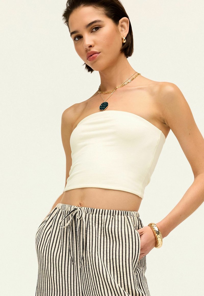 Biały strapless crop top w połączeniu z szarymi i białymi, luźnymi spodniami w paski. Dodatki obejmują warstwowy złoty naszyjnik i bransoletkę.