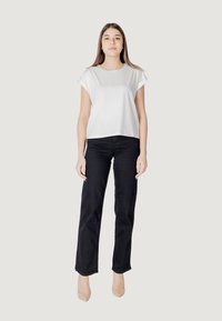 T-shirt bianca corta con maniche arrotolate, abbinata a jeans neri a vita alta e scarpe con tacchi alti color nude, in piedi davanti a uno sfondo neutro.