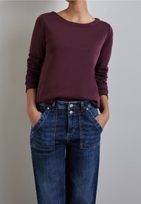 Camisa de manga larga color marrón burdeos con un escote amplio, combinada con jeans de mezclilla azul de cintura alta y de corte holgado, con bolsillos frontales y cierre de botón.