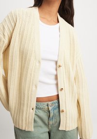 Žena v krémovém pleteném cardigan s knoflíky přes bílý drážkovaný crop top a světle modré džíny s vysokým pasem.