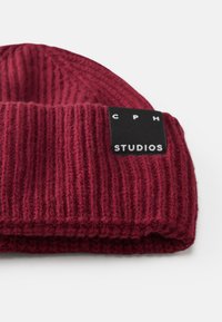 Beanie in magenta lavorato a maglia con texture a costine e etichetta nera con testo bianco "CPH STUDIOS". Materiale morbido, design con risvolto.