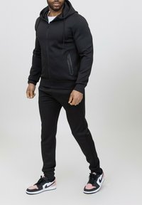 Sweat à capuche zippé noir et pantalon de survêtement assorti en tissu texturé, associés à des baskets noires et roses. Comprend des poches zippées et une coupe ajustée.