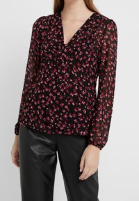 Blouse noire à manches longues avec un motif floral rose, encolure en V et détail froncé à l'avant. Tissu transparent avec poignets élastiques.