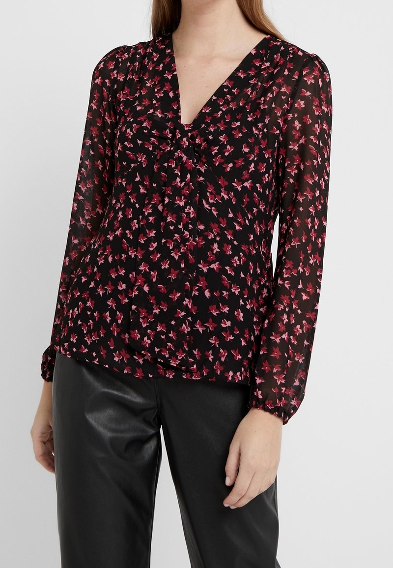 Blouse noire à manches longues avec un motif floral rose, encolure en V et détail froncé à l'avant. Tissu transparent avec poignets élastiques.