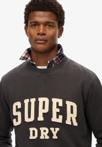 Sudadera gris oscuro con un logo bordado color crema "SUPER DRY." Superpuesta con un cuello de camisa a cuadros en rojo, verde y blanco.