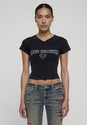 Donna con capelli scuri, in una maglietta corta nera aderente con la scritta "True Religion", indossa jeans blu consumati con toppe di testo rosse.