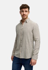 Camicia a righe beige e blu navy, con maniche lunghe, colletto a punta e bottoni in legno. Indossata sopra dei jeans blu.