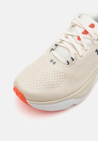 Beige joggesko med mesh-overdel, hvit mellomsåle og kraftig oransje yttersåle. Har sikre snøring og Under Armour-logo på siden.
