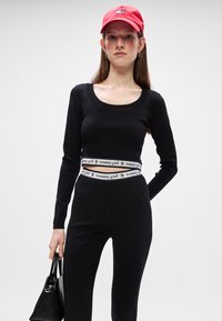 Crna majica dugih rukava s izrezom u obliku uže, u kombinaciji s crnim hlačama. Bijeli brendirani pojas s crvenim zvijezdama i crvena kapa. Crna torba.