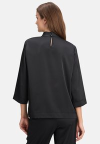 Schwarzes Satin-Oberteil mit drei Viertelärmeln, hohem Ausschnitt und einer Schlüsselloch-Details am Rücken. Glatte Textur, lässiges Design.